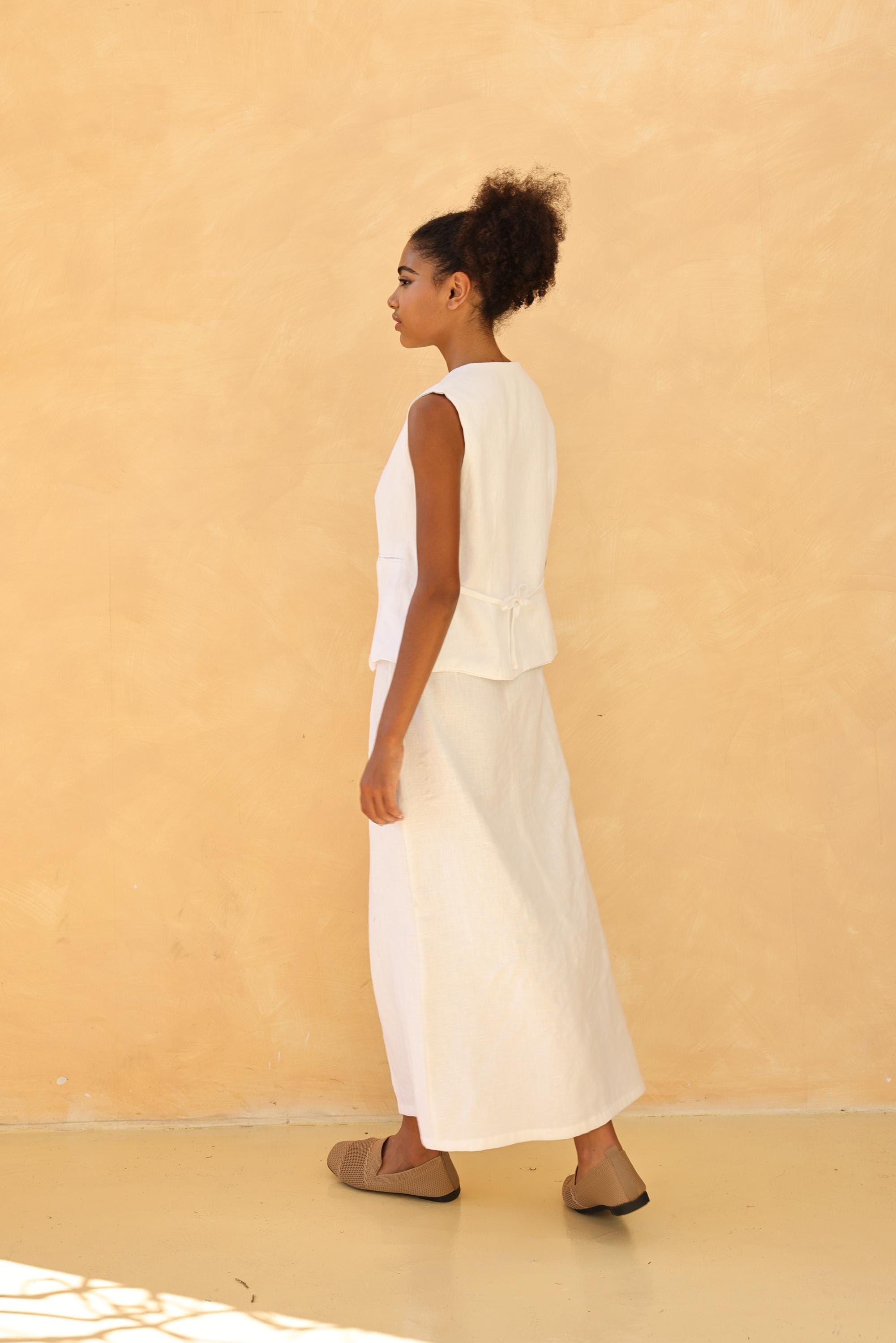 Woman wearing a white sleeveless top and long white skirt against a beige background