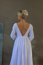 Woman wearing a white wedding dress with a low back and 3/4 sleeves against a plain background