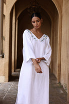 Woman in a white dress with floral embroidery standing in an arched stone corridor.