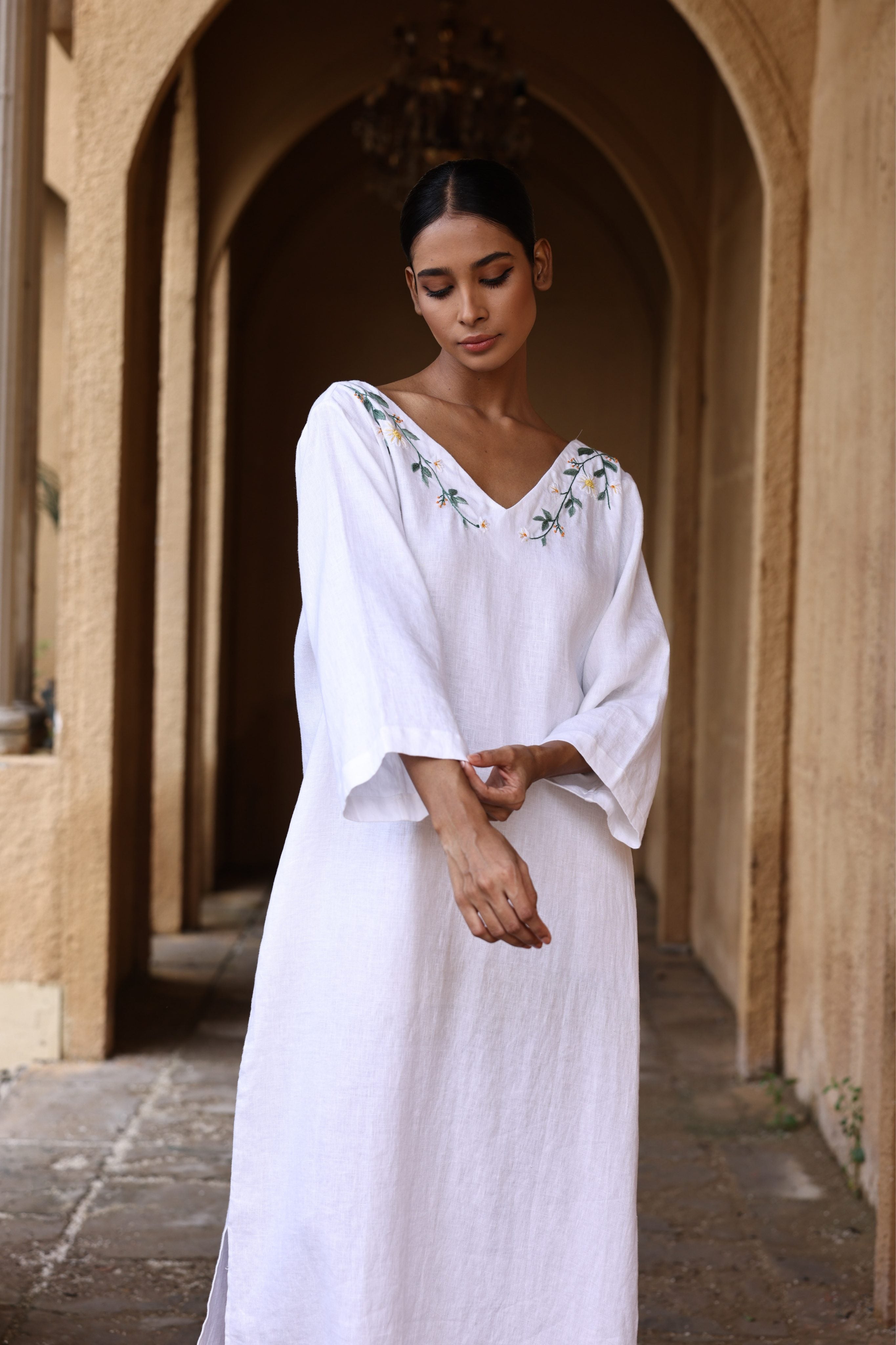Woman in a white dress with floral embroidery standing in an arched stone corridor.