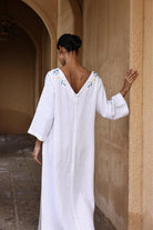 Woman in a white dress with floral embroidery standing against a beige wall.