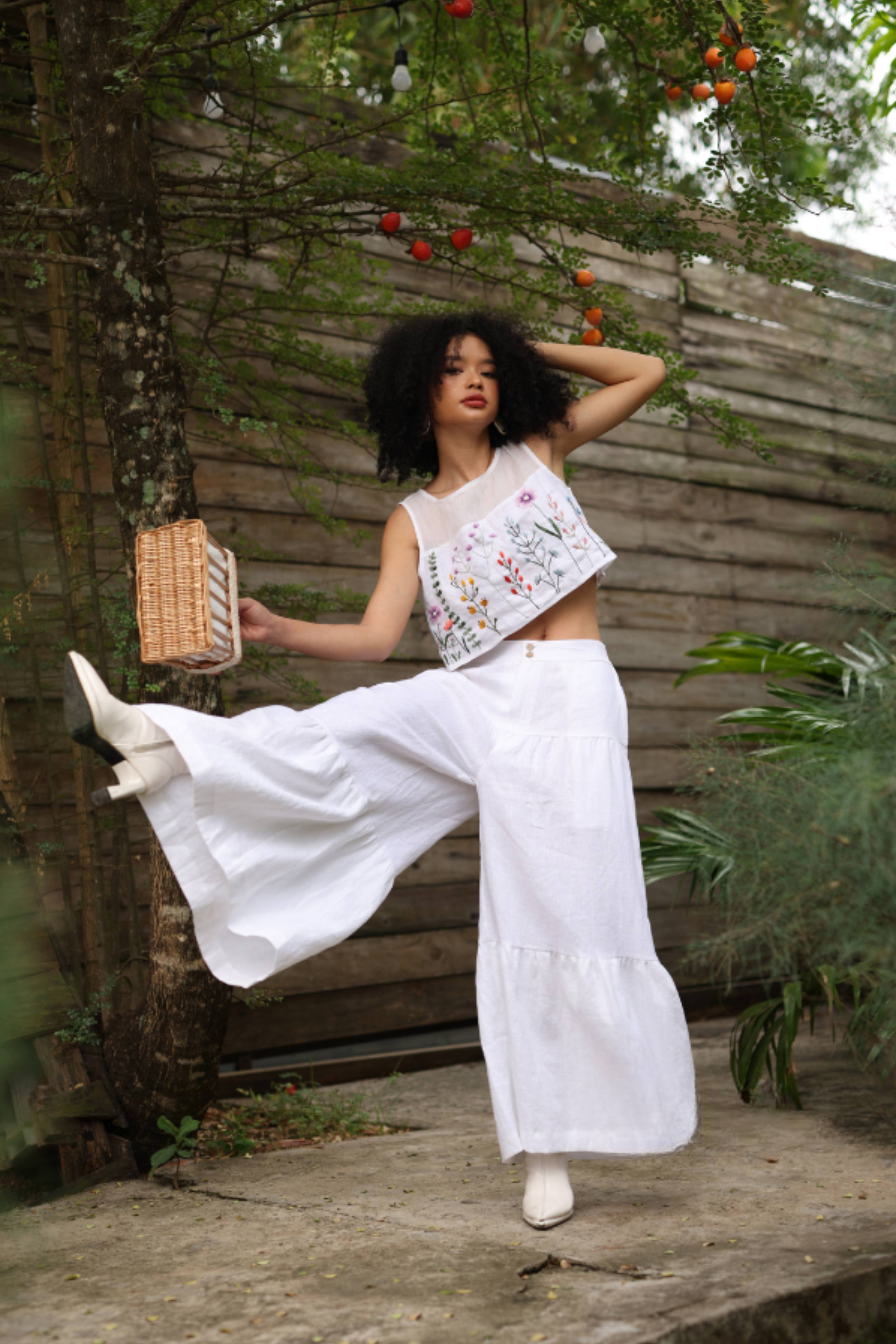 Woman in a white outfit with floral embroidery standing outdoors near a wooden fence and trees.