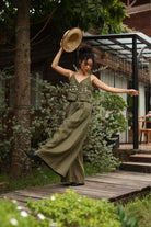 Woman in a green outfit and hat dancing outdoors on a wooden path with greenery and a building in the background.