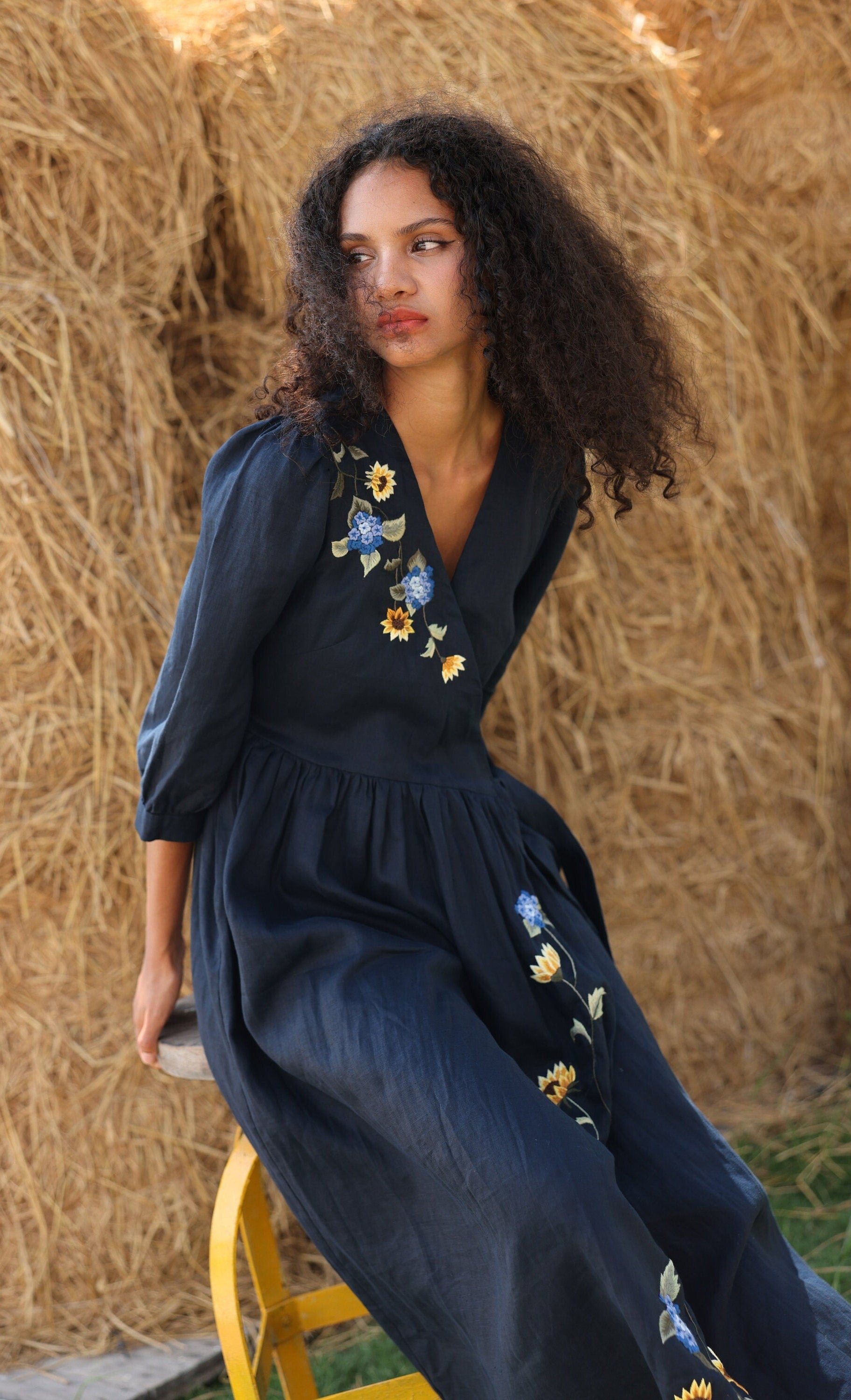 Woman in a dark blue dress with floral embroidery sitting on a yellow stool against a hay bale.